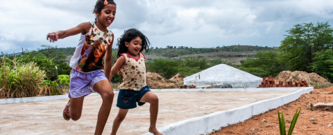 Duas meninas correndo em um terreno e tem uma cisterna ao fundo